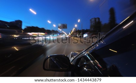 A dark-colored car is moving rapidly along the illuminated street of a night city, on a blurred abstract background a part of the car with a mirror is visible