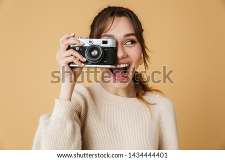Attractive young woman wearing sweater standing isolated over beige background, taking a picture with photo camera