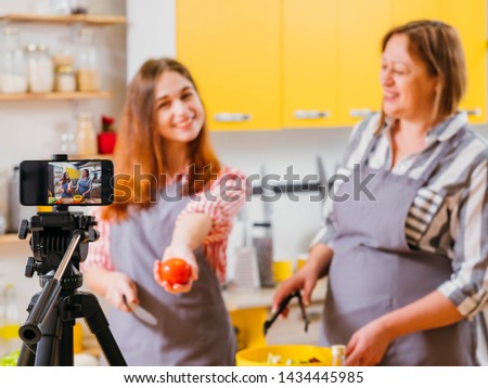 Family culinary video blog. Mother and daughter using smartphone on tripod to film steps of preparing vegetable salad.