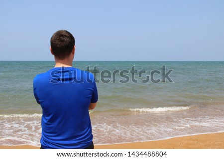 a man stands with his back in a blue t-shirt for swimming against the background of the sea and sand. sunny summer day. the man has dark hair Royalty-Free Stock Photo #1434488804