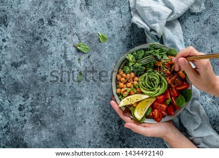 Hands holding healthy superbowl or Buddha bowl with salad, baked sweet potatoes, chickpeas, broccoli, hummus, avocado, sprouts on light blue background. Healthy vegan food, clean eating, top view 