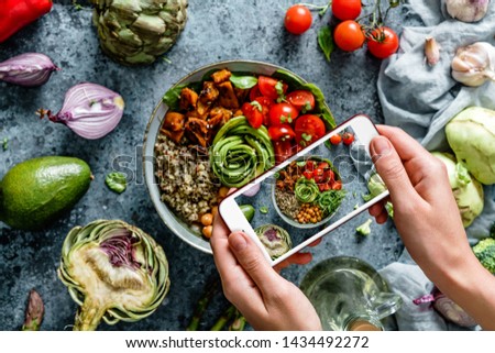 Girl's hands taking photo of Buddha bowl salad with chickpeas, quinoa, tomatoes, arugula, avocado, sprouts by smartphone. Healthy vegan food, clean eating, dieting, top view