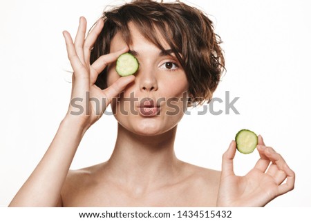 Beauty portrait of a lovely young topless woman with short brunette hair standing isolated over white background, holding sliced cucumber