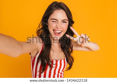 Happy cheerful girl wearing summer outfit standing isolated over yellow background, taking a selfie, showing peace gesture