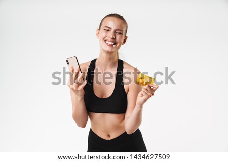 Attractive happy young fit sportswoman standing isolated over white background, using mobile phone and showing credit card