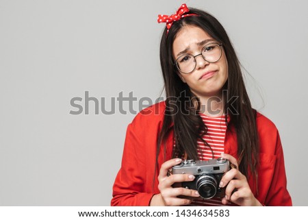 Cute upset teenage girl wearing casual outfit standing isolated over gray background, holding photo camera