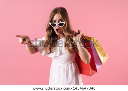 Beautiful shocked young blonde girl wearing summer dress standing isolated over pink background, carrying shopping bags, pointing away