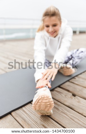 Attractive smiling young sportswoman wearing hoodie sitting on a fitness mat outdoors at the beach, stretching legs