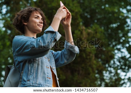 Attractive young girl wearing casual outfit spending time outdoors at the park, taking pictures with mobile phone