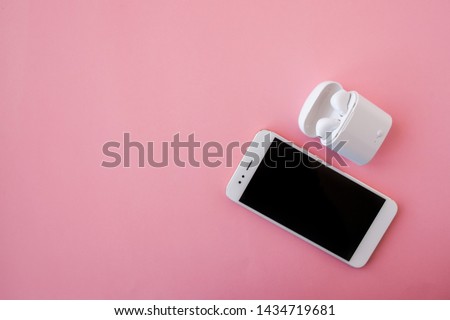 White wireless headphones and smartphone are lying on a bright pink background. Headphones in charging case. Top view. Flatlay, Copy space left.