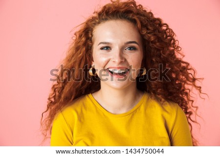 Close up portrait of an attractive cheerful young woman with long curly red hair standing isolated over pink background