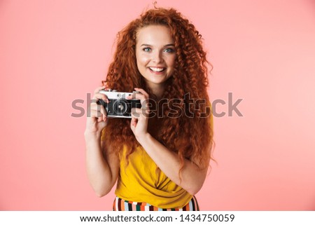 Portrait of an attractive cheerful young woman with long curly red hair standing isolated over pink background, taking pictures with photo camera