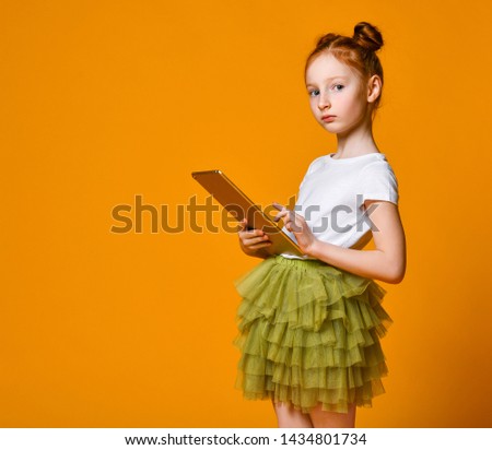 Cute red-haired girl school girl holding a tablet and looks into it. Side view. Child playing game, surfing or watching cartoon