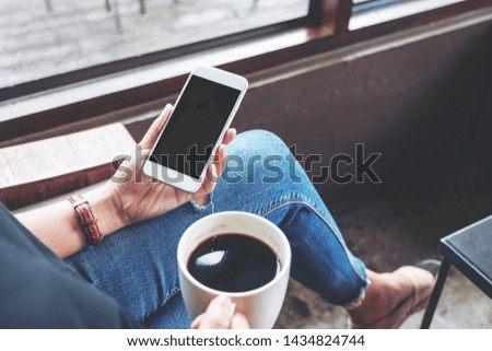 Mockup image of a woman holding white mobile phone with blank desktop screen while sitting and drinking coffee in cafe
