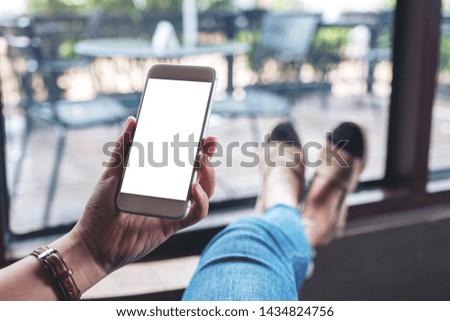 Mockup image of a woman holding white mobile phone with blank desktop screen while sitting in cafe