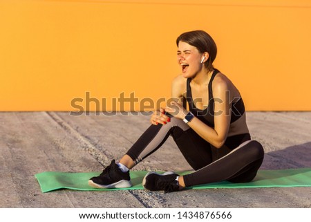 Young sporty girl in black sportwear sitting on mat and have a strong hurt problem with knee, spasm painful. Gripping leg with hand and screaming. Outdoor shot. Sport injury healthy medical concept