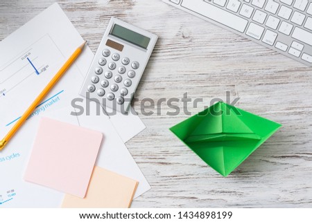 Modern office workspace with green paper ship. Eco-friendly business idea. Flat lay wooden desk with computer keyboard, calculator and documents. Creative marketing and advertising agency.