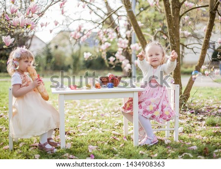 Two cute little blonde girls of 3 years old are playing in the park near a blossoming magnolia. Tea drinking. Easter. Spring.