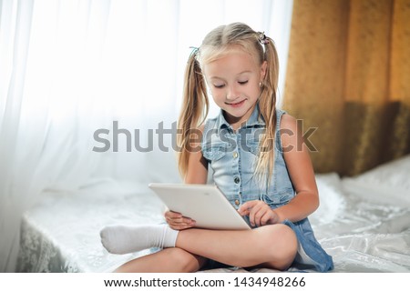 Cute little child girl blond in denim sundress lies in bed uses digital tablet. child playing on tablet pc having fun and smiles