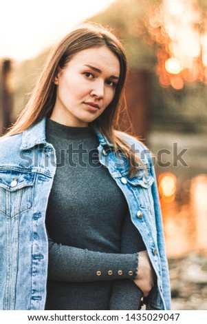 Portrait of the attractive, slender, beautiful young Caucasian blonde girl in a jeans jacket. Smiling girl enjoys fine warm sunner weather against the forest