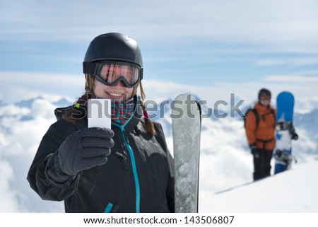 Attractive girl holding blank lift pass and ski smiling with snowboarder and mountain range in background. Concept to illustrate ski admission fee