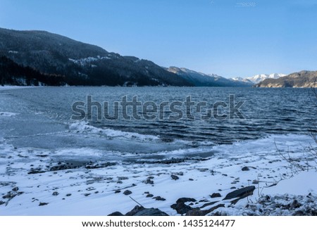 Beautiful winter landscape, snowy mountains at Harrison Hot Springs, British Columbia, Canada. Blue sky. Blue Harrison lake. Frozen ice on rocks in the foreground. Cold, freezing weather. Trees
