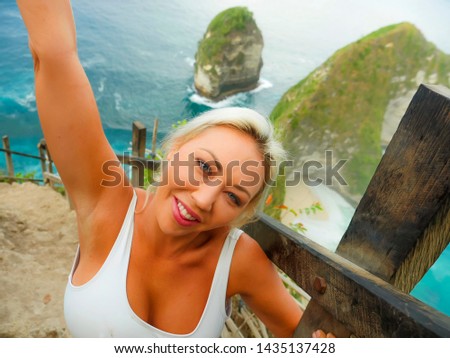lifestyle portrait of young attractive and happy blond tourist woman taking selfie at beautiful sea cliff beach viewpoint enjoying exotic holidays adventure in tropical paradise island