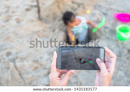 Close up of mother's hand taking photo of baby boy playing sand at beach with smartphone.