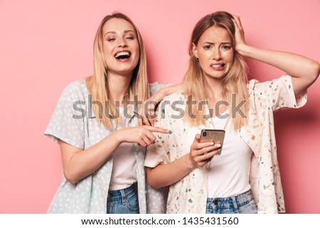 Two attractive blonde girls wearing summer outfit standing isolated over pink background, confused girl holding mobile phone