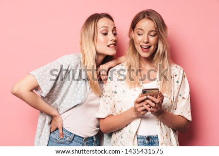Two attractive blonde girls wearing summer outfit standing isolated over pink background, using mobile phone