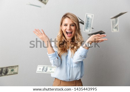 Attractive young blonde businesswoman wearing shirt standing under money banknotes rain isolated over gray background