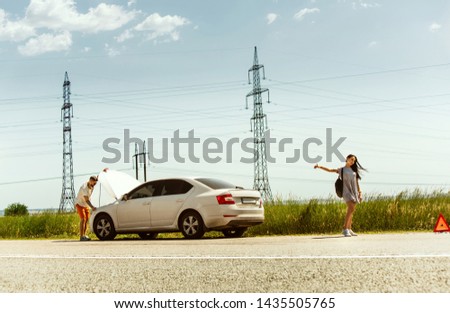 The young couple broke down the car while traveling on the way to rest. They are trying to stop other drivers and ask for help or hitchhike. Relationship, troubles on the road, vacation.