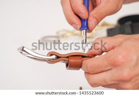 man makes by hands leather belt with buckle. handmade hobby. young man resting by manufacture his designer belts. man with a screwdriver in his hands twists rivets close up on white studio background.