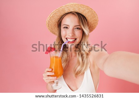 Happy pretty young girl wearing swimsuit standing isolated over pink background, taking a selfie, holding a cocktail