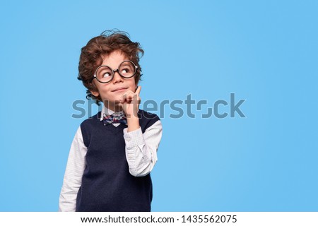 Nerdy boy in round eyeglasses and school uniform rubbing chin and looking away while thinking near empty space against blue background