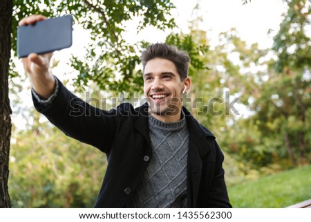 Image of cheerful young man in casual clothing walking outdoors in green park using mobile phone take a selfie.