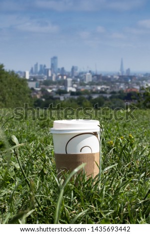 A picture presenting a cup of coffee put on a grass in front of the famous London's view spot: the skyline of London seen from Hampstead Heath.