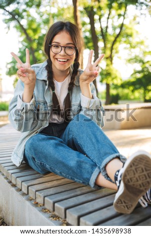 Photo of a cheerful cute young student girl wearing eyeglasses sitting on bench outdoors in nature park make peace gesture.