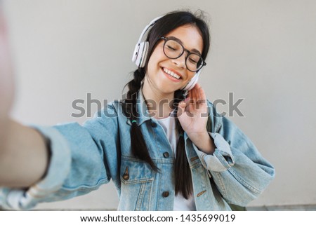 Portrait of a pleased cute beautiful girl in denim jacket wearing eyeglasses isolated over gray background listening music with headphones take a selfie by camera.
