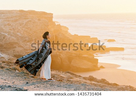 Young woman with flying blanket standing on cliff's edge and looking to the ocean on sunset. Wanderlust concept. Royalty-Free Stock Photo #1435731404