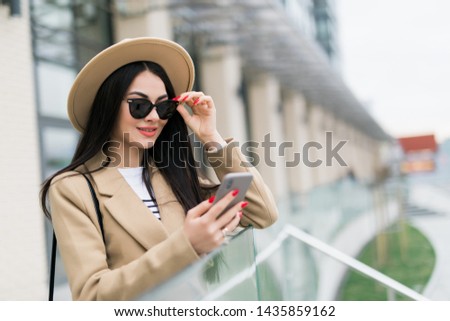 Beautiful girl in a beige coat, holds a smartphone in her hands, stands in front of the building's urban background, looks at the phone screen.