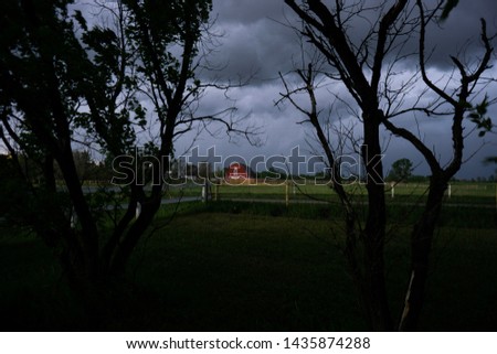 Dark and stormy night with a red barn in the middle of a field framed by dark waving trees