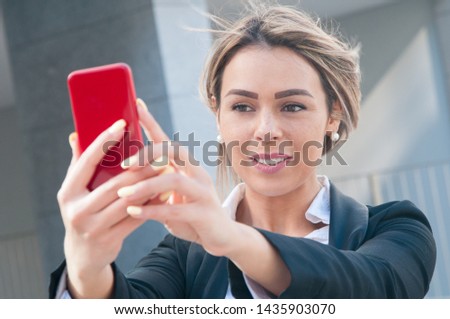 Smiling business lady talking selfie photo outdoors. Pretty young woman standing and posing at smartphone camera with blurred building view in background. Selfie and business concept. Front view.