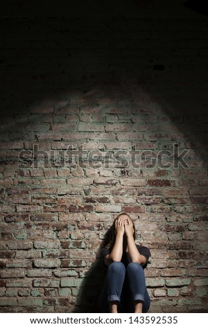 Depressed woman sitting alone against brick wall with light coming from above.