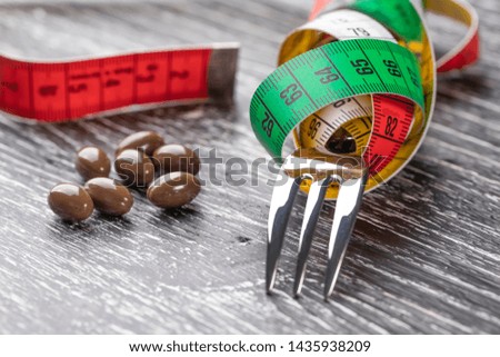 A shiny fork wrapped in a multi-colored centimeter to measure body shapes and brown medical capsules on a black old wooden table. The concept of nutritional supplements for weight loss