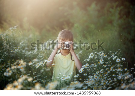 Cute boy photographer shoots on camera in nature. Kid takes a photo in the camomile flowers field on sunset summer.