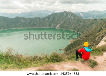 Woman hiker in red looking at dramatic perspective of Quilotoa lake and volcano crater, with view of mountains, hiking loop from viewpoint. Shot in Ecuador. Green and blue. Freedom, adventure.