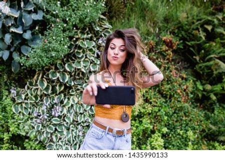 young blonde beautiful woman at the street taking a picture with mobile phone and smiling. Lifestyle outdoors. Summertime, Green background