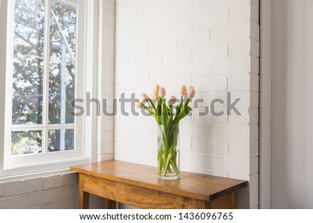 Pale pink tulips in glass vase on wooden sidetable against white painted brick wall and window (selective focus)