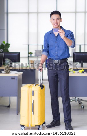 Young man stand in his office looking at the camera and smile, left hand's showing credit card and right hand holding big yellow luggage. Prepare for the upcoming holiday. Holiday concept